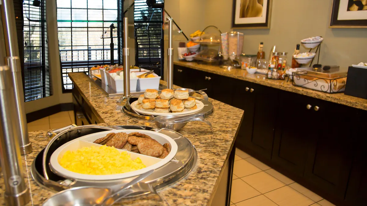 A bright, sociable dining area features a granite countertop breakfast buffet, surrounded by ample seating for guests to enjoy meals together. The atmosphere is friendly and lively, with natural light streaming in and guests connecting over food.