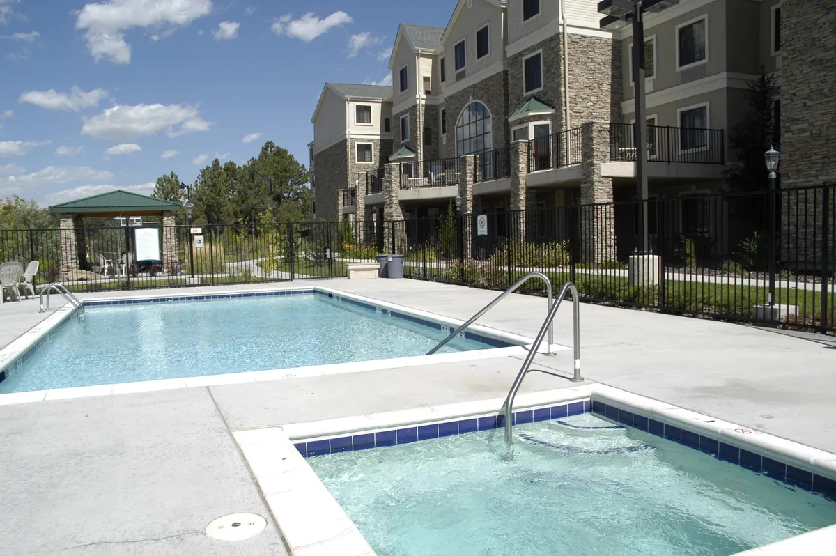 This image showcases the inviting outdoor swimming pool and adjacent hot tub, surrounded by lush landscaping and the building exterior. Guests can be seen enjoying both swimming and relaxing in the hot tub, evoking a tranquil, resort-style atmosphere that highlights both seasonal and year-round amenities.