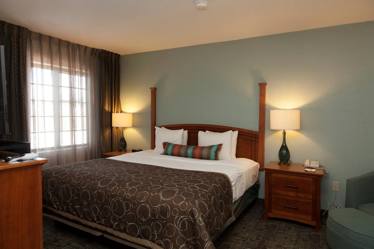 This image shows a serene bedroom featuring a king-sized bed adorned with decorative pillows, flanked by nightstands and softly glowing lamps. A large window dressed with curtains allows natural light to brighten the space, and a TV unit offers entertainment. The overall feel is cozy and inviting, ideal for rest and relaxation.