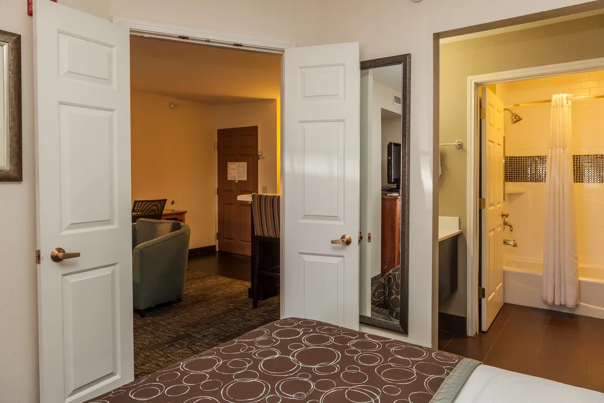 A cozy interior bedroom scene showing a bed with an open doorway, a mirror reflecting part of the room, and a comfortable seating area. The space feels intimate, designed for quiet moments or shared conversations after a busy day.