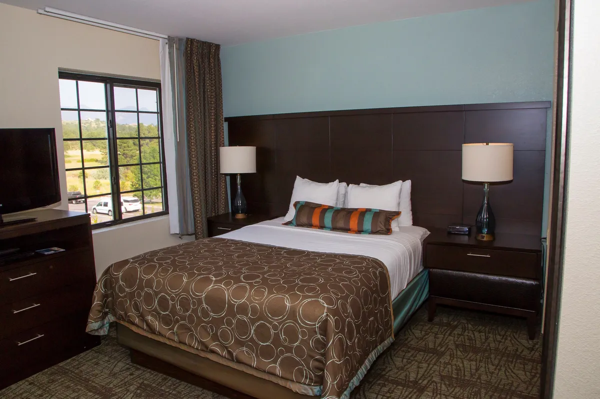 This image features a bedroom with a queen-sized bed topped with decorative pillows, a nightstand with a lamp, and a television across from the bed. The ambiance is peaceful and softly lit, with morning sunlight streaming in, creating a restful space for unwinding.