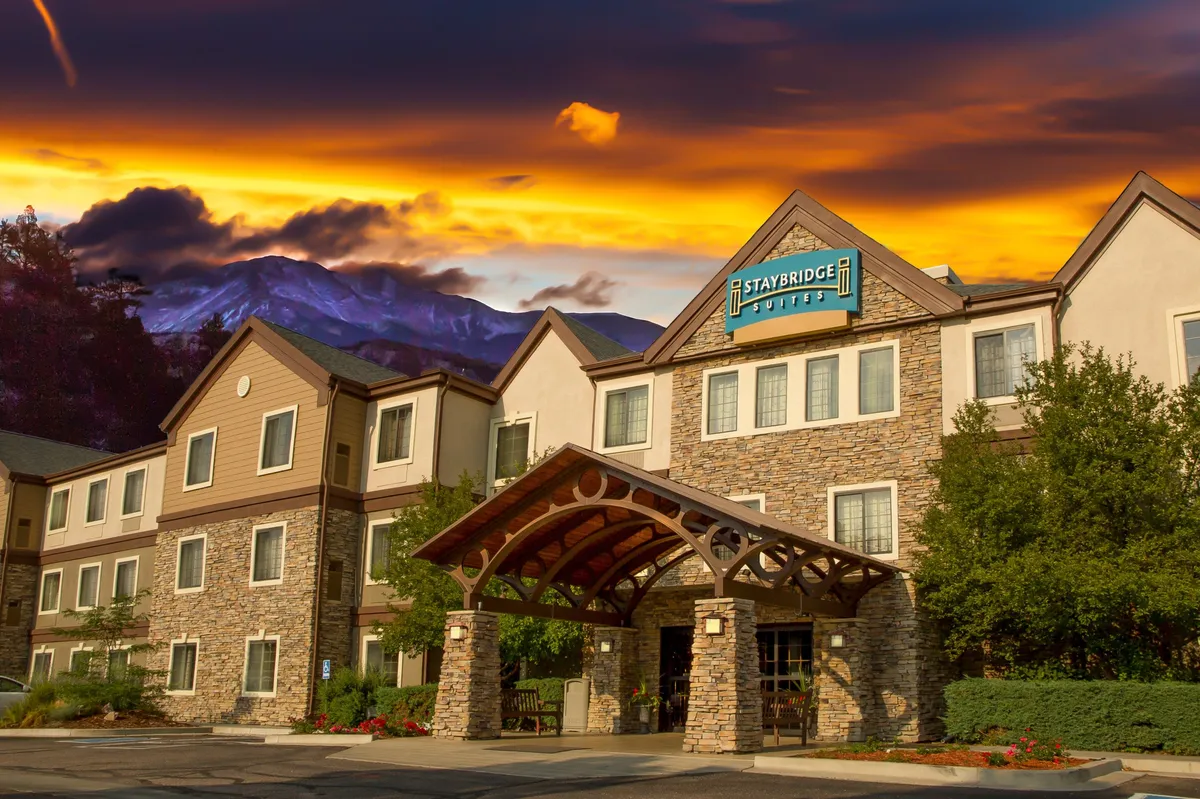 This image shows the resort's exterior, featuring a stone facade, a charming wooden awning, landscaped grounds, and a scenic mountain backdrop. The outdoor setup suggests a welcoming environment for family gatherings and meals outdoors, strongly evoking the experience of grilling or dining al fresco.